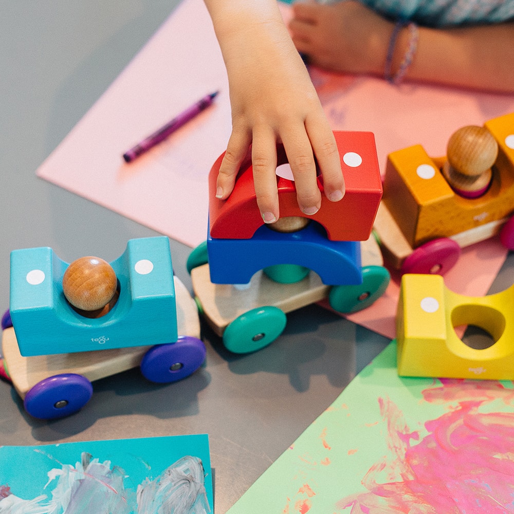 The image shows the hands of a child playing the toys, on colored sheets of paper with pink and white paint on them.